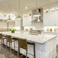 White kitchen design features large bar style kitchen island with granite countertop illuminated by modern pendant lights. Stainless steel appliances framed by white shaker cabinets . Northwest USA